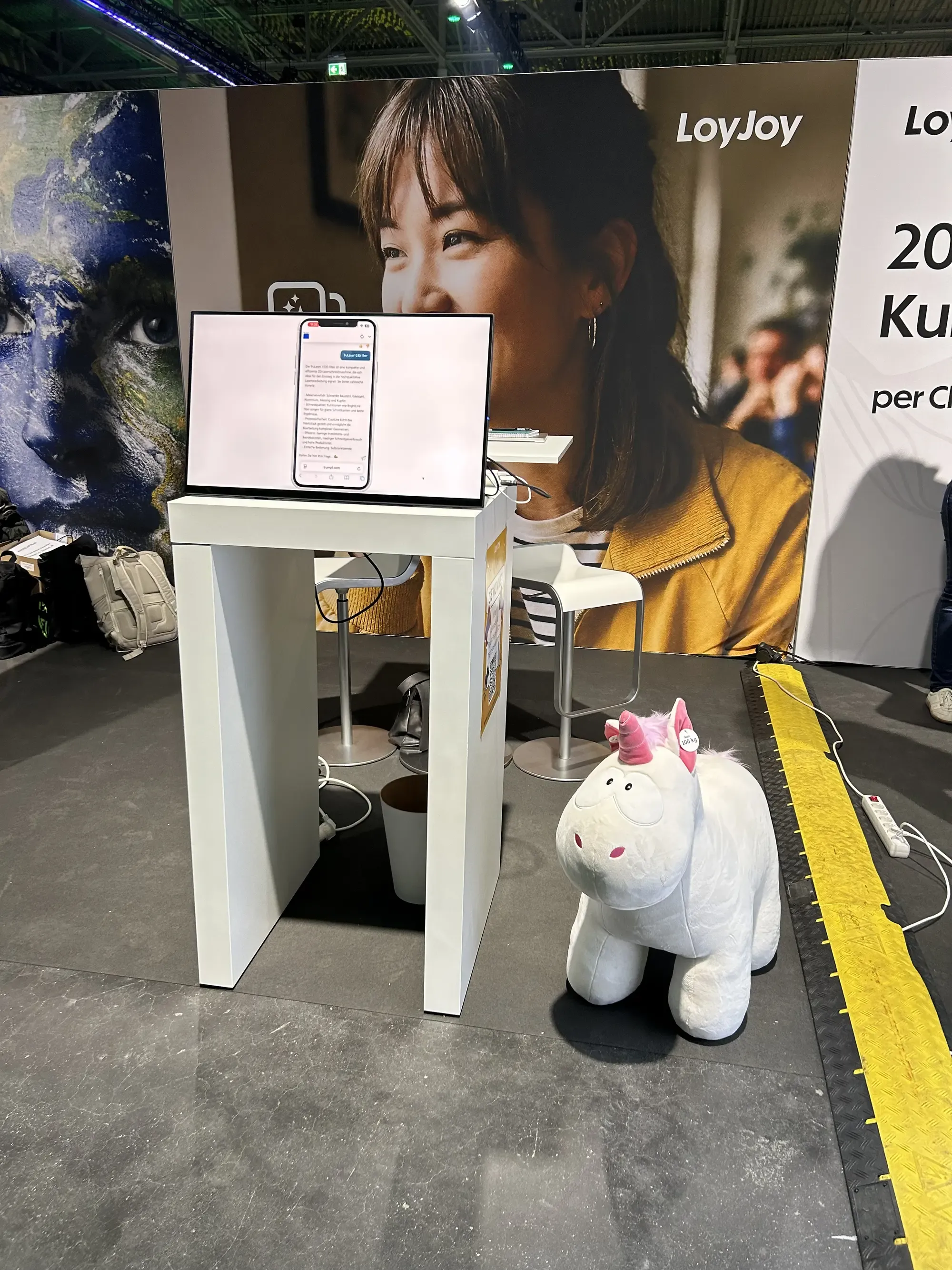 Man sieht einen Teil unseres Standes. Auf einem Tisch steht ein Bildschirm, an dem man LoyJoy ausprobieren kann. Daneben steht ein großes Einhorn, was im Gewinnspiel verlost wurde. Im Hintergrund sieht man noch ein Banner mit dem LoyJoy Logo drauf.