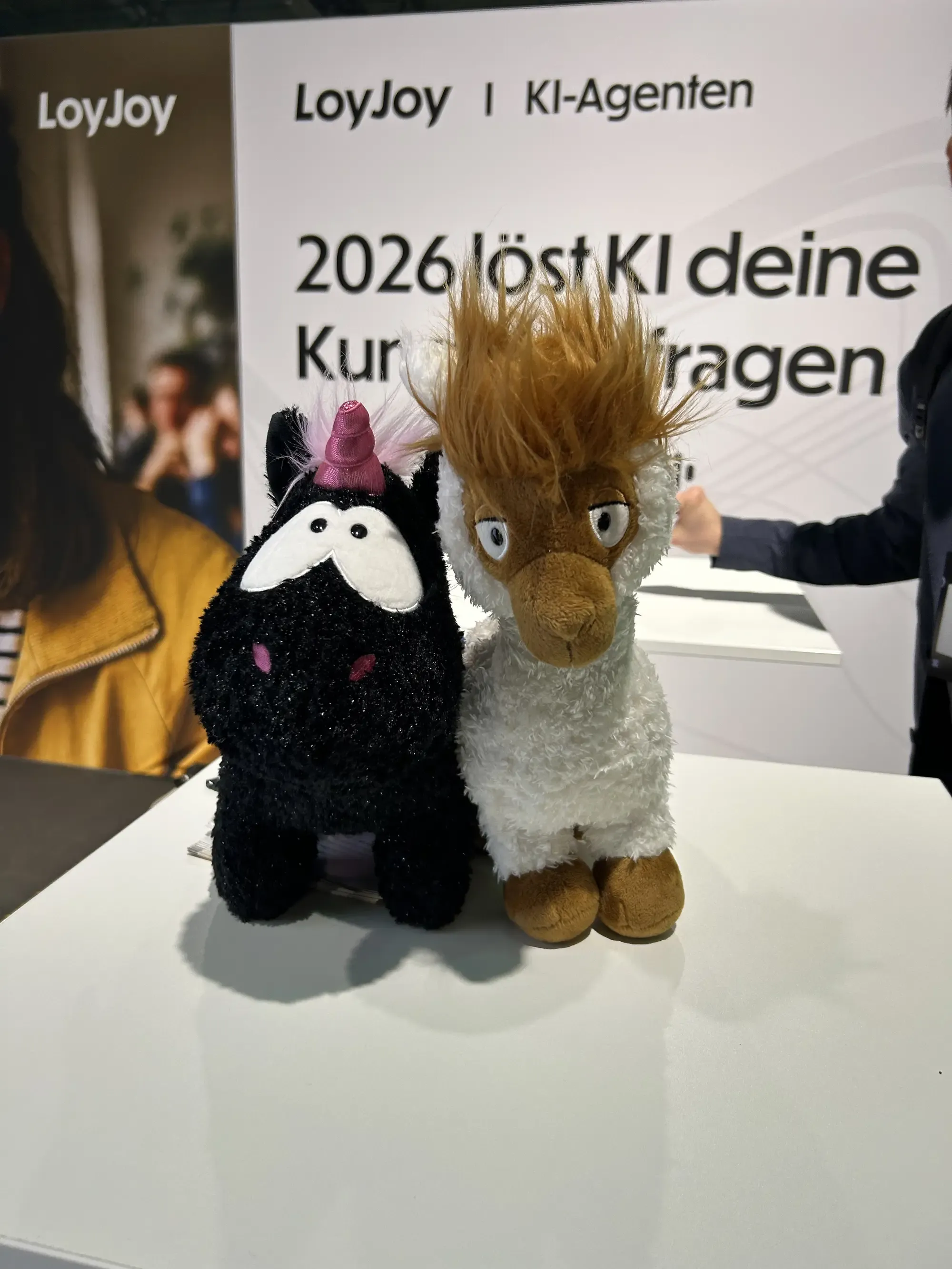 Ein Einhorn und ein Alpaka aus unserem Gewinnspiel stehen auf einem Tisch an unserem Stand. Im Hintergrund sieht man ein Banner mit dem LoyJoy Logo.