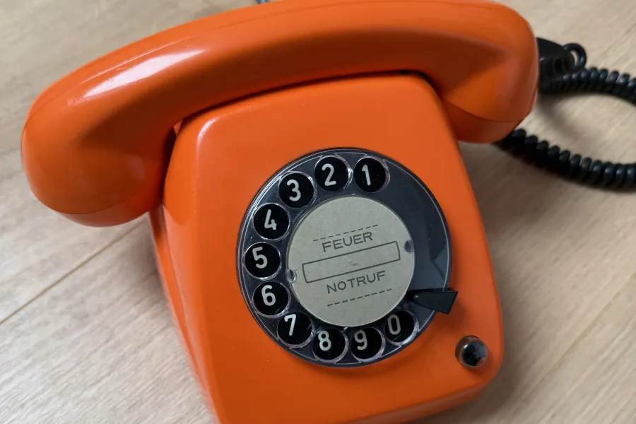 Orangefarbenes Wählscheibentelefon FeTAp 615 mit Notruf-Etikett auf einem Holzboden