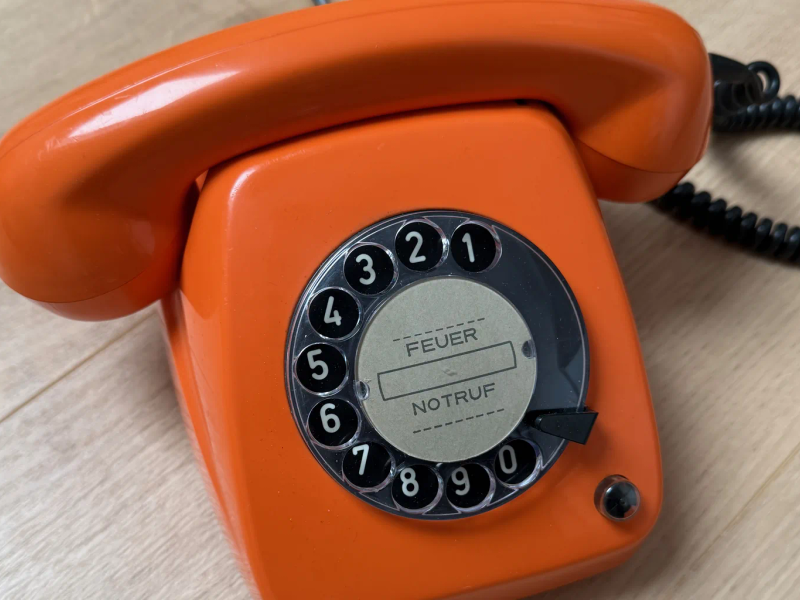 Orangefarbenes Wählscheibentelefon FeTAp 615 mit Notruf-Etikett auf einem Holzboden