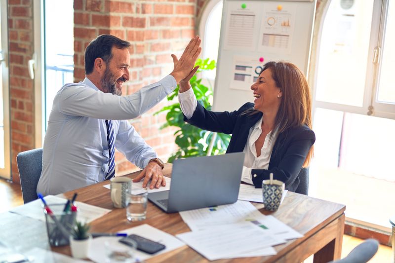 Two people in an office are smiling and exchanging a high-five with each other.