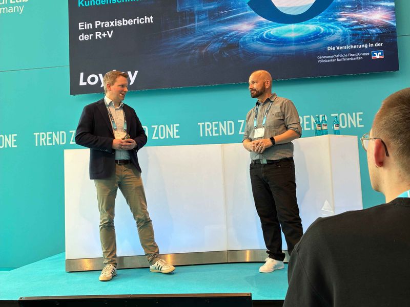 Ulf Loetschert from LoyJoy and Peter Paul Nasim from R+V Versicherung are giving a joint presentation on stage at insureNXT in the Trend Zone. In the background, a large screen displays the presentation entitled ‘Agentic AI at the customer interface’.